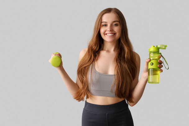 Sportliche junge Frau mit Wasserflasche und Hantel auf grauem Hintergrund - Foto, Bild