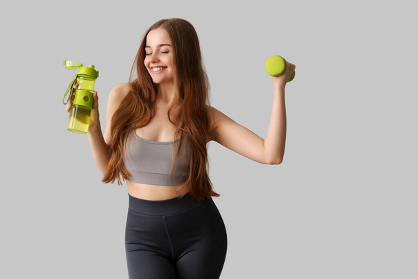 Sporty young woman with bottle of water and dumbbell on grey background - Фото, изображение