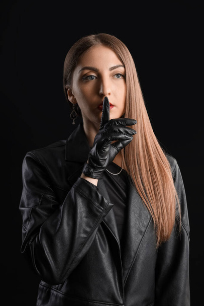 Beautiful young woman in leather gloves and jacket showing silence gesture on black background - Φωτογραφία, εικόνα