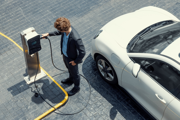Aerial view of progressive businessman in black formal suit with his electric vehicle recharging battery at public car park charging station as vehicle powered by sustainable energy concept. - Photo, image