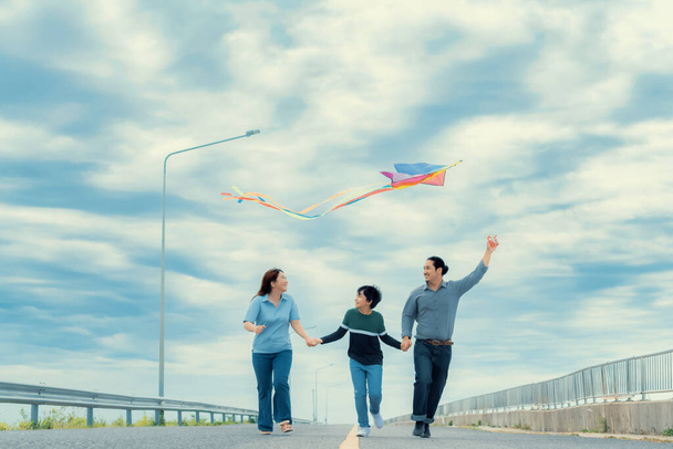 Progressive happy family vacation and carefree day concept. Young parents mother father and son run along and flying kite together road with enjoy natural scenic on scenery and clear sky background. - Fotoğraf, Görsel