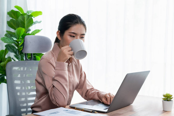 Jonge Aziatische enthousiaste zakenvrouw werken aan moderne bureau, drinken van koffie als ze ijverig werken op haar laptop vertegenwoordigen professionele en aantrekkelijke kantoormedewerker in haar werkruimte. - Foto, afbeelding