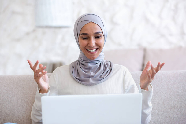 Close-up photo. Muslim young woman in light hijab talking on laptop video call at home. He smiles, greets, gestures with his hands. - Photo, Image