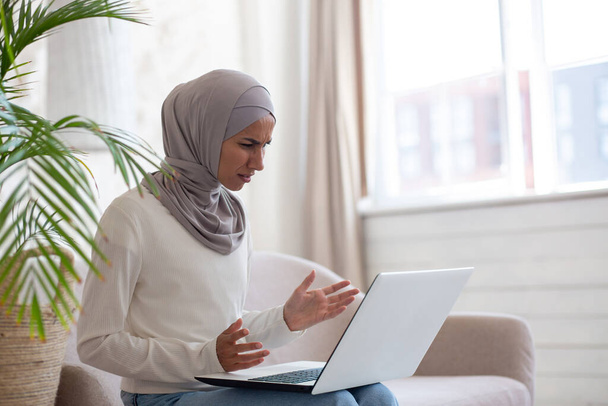 Arab young woman in hijab sitting on sofa at home, holding laptop on lap, looking worriedly at screen and spreading hands. There are problems, she is stressed, upset. - Photo, image