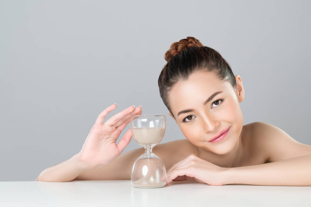 Glamorous model holding hourglass in beauty concept of anti-aging skincare treatment for woman. Young asian girl portrait with perfect smooth clean skin and flawless soft makeup in isolated background - Foto, Bild