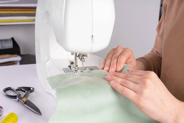 Las manos femeninas que cosen la tela blanca en la máquina de coser moderna en el lugar de trabajo en el taller. Las manos de las mujeres cosen piezas de tela en un primer plano de la máquina de coser. Hecho a mano, hobby, concepto de pequeña empresa - Foto, Imagen