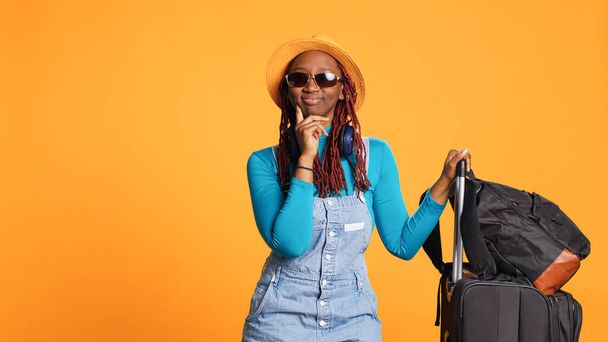 Ενθουσιασμένη γυναίκα που φοράει γυαλιά ηλίου και καπέλο, αισθάνεται σιγουριά ταξιδεύοντας στον προορισμό των διακοπών. Νεαρός ενήλικας με αποσκευές και τσάντες τρόλεϊ που αναχωρούν για αστική περιπέτεια, δροσερά γυαλιά. - Φωτογραφία, εικόνα