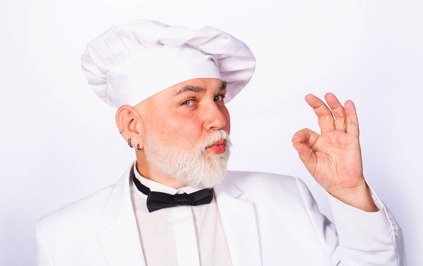 Шеф-повар в белой форме показывает знак ОК. Бородатый повар в шляпе шеф-повара с жестом для утверждения ресторанного обслуживания. Профессиональный повар показывает знак "вкусно". Бородатый повар отлично жестикулирует - Фото, изображение