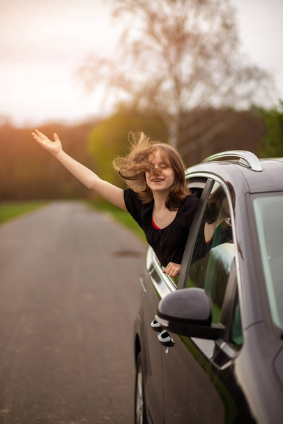 una chica feliz con su pelo revoloteando en el viento mira por la ventana del coche en un día de verano. - Foto, imagen