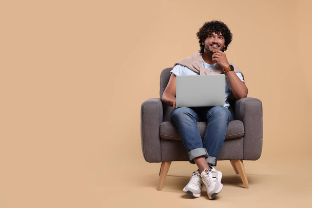 Smiling man with laptop sitting in armchair on beige background, space for text - Φωτογραφία, εικόνα