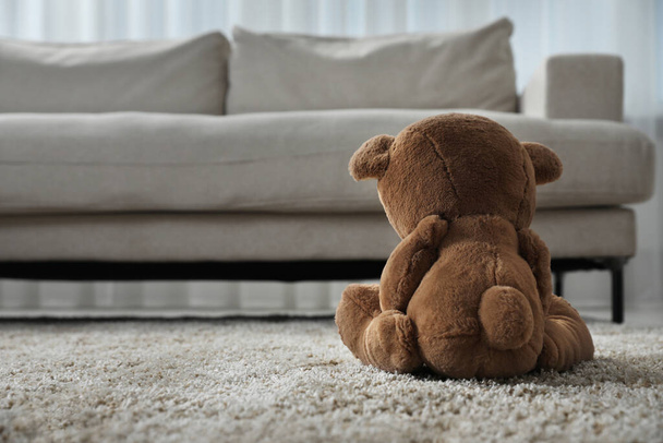 Cute lonely teddy bear on floor near sofa in room, back view. Space for text - Foto, afbeelding