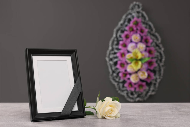 Photo frame with black ribbon, rose on light table and wreath of flowers near grey wall indoors, space for text. Funeral attributes - Φωτογραφία, εικόνα