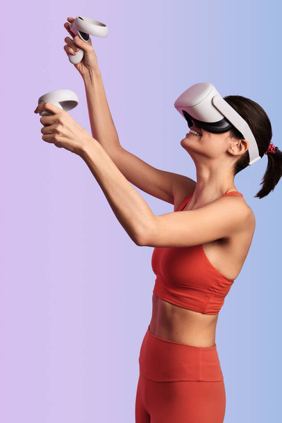 Side view of young smiling female in red sportswear and VR headset using controllers while raising arms and interacting with cyberspace against gradient background - Foto, Imagen