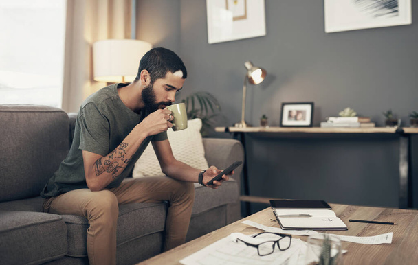 Отслеживаю его расходы с помощью умных приложений. молодой человек, пьющий кофе и пользующийся смартфоном во время бумажной работы дома - Фото, изображение