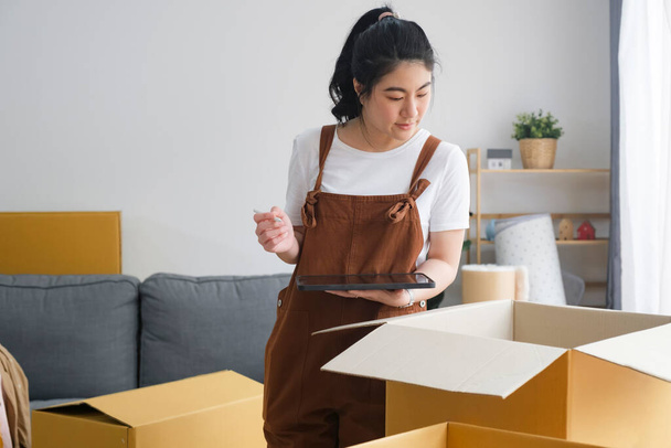 Karton kutuları kontrol eden ve paketleyen Asyalı kadın, kontrol listesiyle çevrimiçi alışveriş yapan müşterilere teslim edilmeden önce yeni ev taşıma, nakliye veya ürün paketlerini kontrol etmeye hazır olun. - Fotoğraf, Görsel