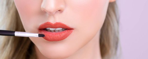 Closeup beautiful young woman with flawless healthy skin and natural makeup putting alluring fashion glossy red lipstick on her lip with lip brush in pink isolated background. - Fotografie, Obrázek