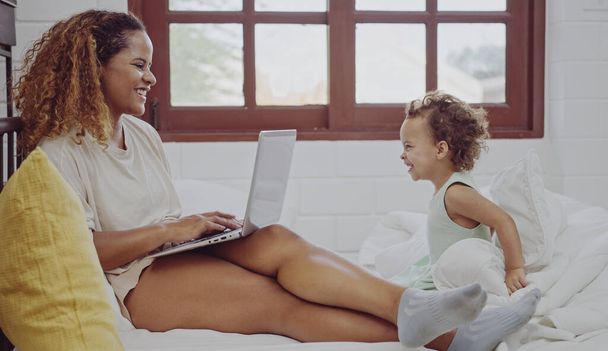 Jonge moeder met behulp van laptop voor werk in de buurt spelen dochter op bed. - Foto, afbeelding