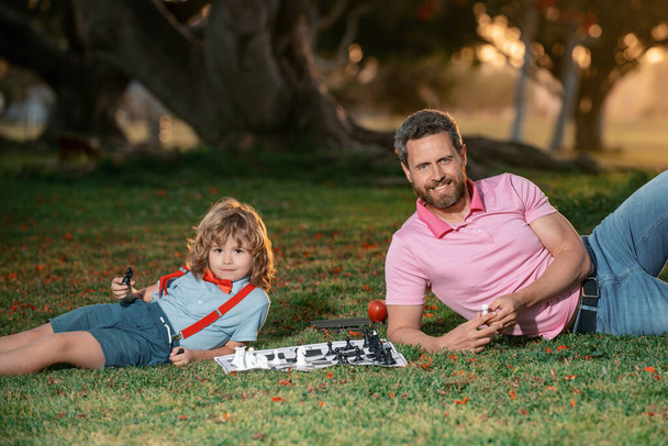 Son laying on grass and playing chess with father. Games and entertainment for children. Family concept - Foto, Imagen