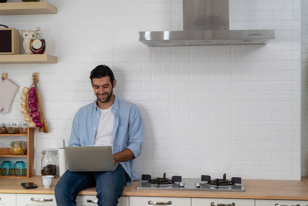 Freelancer working from home sitting on a kitchen worktop and using laptop. Bearded man working with a laptop and reading news. Handsome successful self entrepreneur working at his modern home. - 写真・画像
