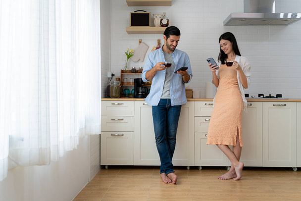Handsome bearded young man and beautiful young woman standing in kitchen drinking morning coffee using mobile smartphone looking for various services and social media on the internet. copy space - Φωτογραφία, εικόνα