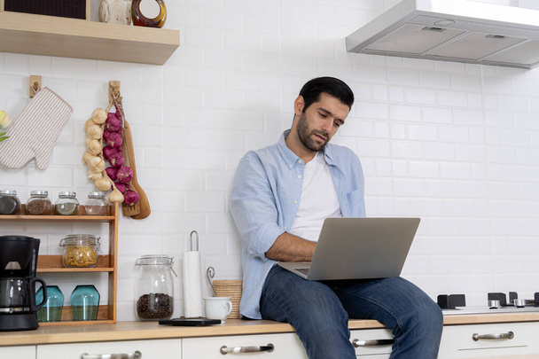 Young handsome businessman struggling to work at his modern home. Freelancers work from home sitting on the kitchen table and using their laptops. Bearded man working with laptop and reading news.  - 写真・画像