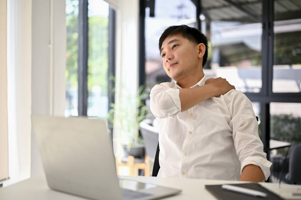 Tired and overworked millennial Asian businessman rubbing his shoulder while sitting at his desk in the office. Office syndrome concept - Foto, Bild