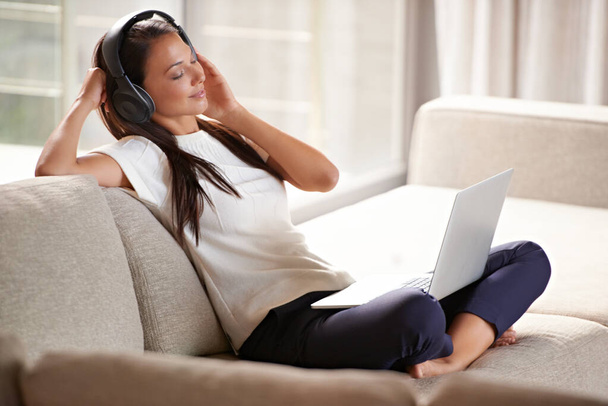 Koptelefoon, laptop en vrouw op een thuisbank luisteren naar muziek of audio tijdens het streamen online. Rustige vrouwelijke persoon ontspannen op de bank om te luisteren naar radio of lied met internetverbinding en technologie. - Foto, afbeelding