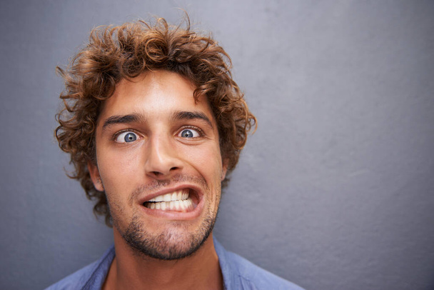 Young man, portrait and silly face for funny or goofy expression against a gray wall background. Male with crazy humor or impression looking and posing in playful happiness or fun manner and attitude. - Foto, Imagen