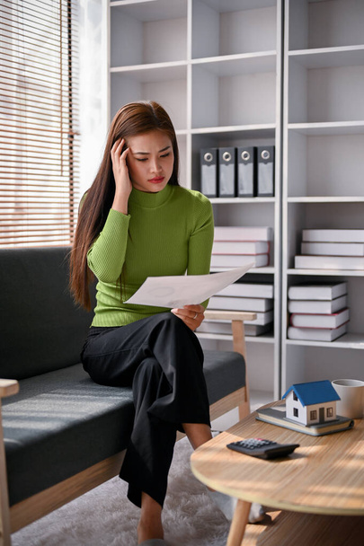 Mujer asiática seria y estresada revisando sus cuentas de gastos mensuales en una sala de estar. - Foto, imagen