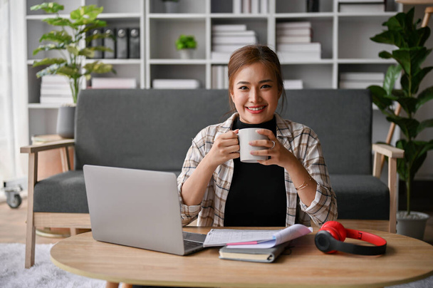 Attractive and smiling young Asian female freelancer sits at a coffee table in her living room with a coffee cup, laptop and stuff. work from home concept - 写真・画像
