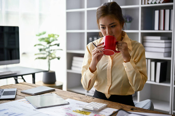 Gericht jonge Aziatische vrouwelijke accountant nippen koffie tijdens het werken aan business financial accounting rapporten aan haar bureau in het kantoor. - Foto, afbeelding