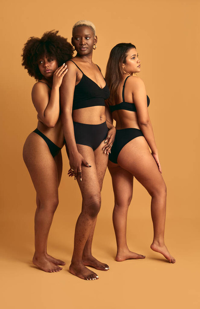 Side view full body of self assured young barefooted multiethnic plus size woman in black underwear standing together against beige background - 写真・画像