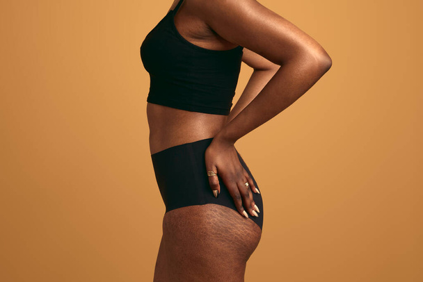 Side view of crop anonymous plus size African woman in black underwear touching buttocks with hands having stretch marks standing against orange background - Foto, imagen