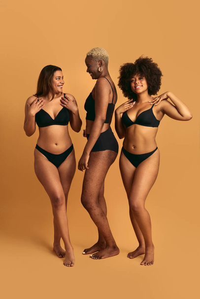 Full length of cheerful confident young multiracial overweight female best friends in black underwear, smiling and looking at each other against beige background in studio - Foto, Imagem