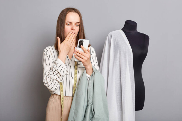 Shocked surprised scared professional female seamstress with mobile phone in hands covering mouth with hand wrong order dressmaking mistake. - Фото, изображение