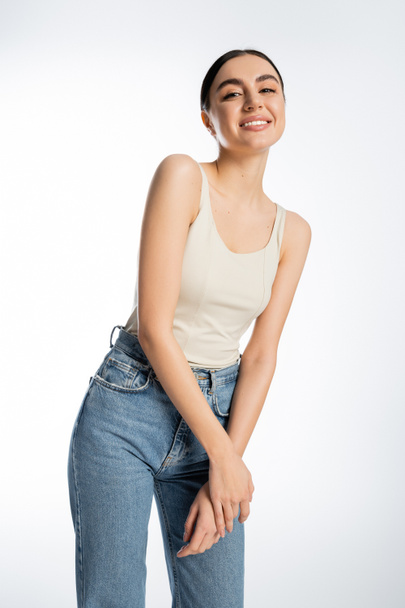 lovely and smiling woman with brunette hair, natural beauty and perfect skin looking at camera while posing in tank top and denim jeans isolated on white background   - Foto, Bild