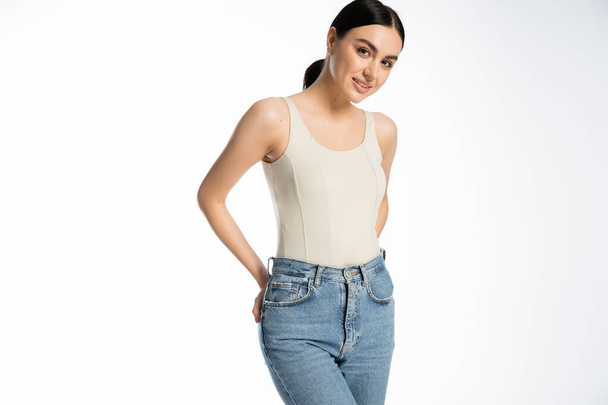 charming and young woman with natural makeup, brunette hair and perfect skin smiling while posing in tank top and denim jeans and looking at camera on white background  - Foto, Bild