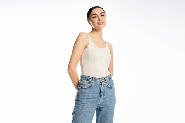 appealing and young woman with natural makeup, brunette hair and perfect skin smiling while posing in tank top and denim jeans and looking at camera on white background  - Foto, Imagem