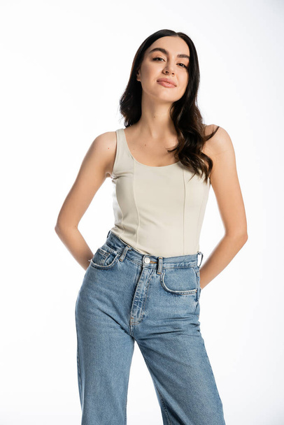 charming woman with natural beauty, brunette hair and perfect skin smiling while posing in denim jeans and tank top and looking at camera on white background  - Foto, imagen