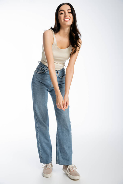 full length of enchanting woman with brunette hair, perfect skin and natural makeup standing in denim jeans and tank top while smiling and looking at camera on white background  - Foto, Bild