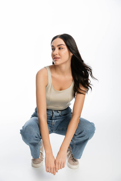 full length of young and carefree woman with natural makeup, brunette hair and perfect skin sitting in denim jeans and tank top and looking away on white background  - 写真・画像