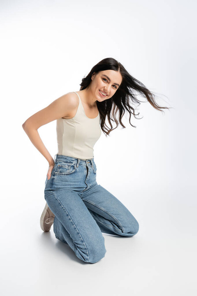 full length of young and gorgeous woman with flawless makeup, brunette hair and perfect skin standing on knees and posing in denim jeans with tank top while on white background  - Foto, afbeelding