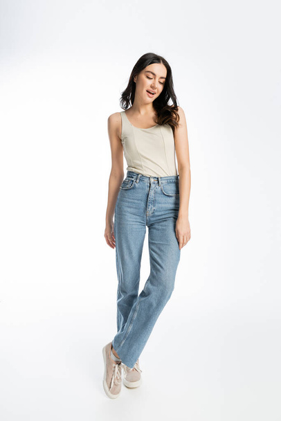 full length of alluring woman with opened mouth, natural makeup, brunette hair and perfect skin posing in tank top and denim jeans on white background - 写真・画像