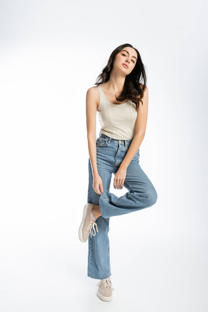 full length of alluring woman with shiny brunette hair, natural makeup and perfect skin posing in tank top and touching denim jeans on white background - Фото, зображення