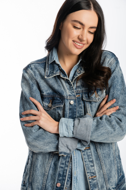 happy and young woman with gorgeous brunette hair posing with folded arms while standing in blue denim jacket and looking down on white background  - Φωτογραφία, εικόνα