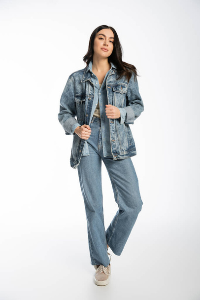 full length of alluring young model with brunette hair, flawless and natural makeup posing in denim jacket and blue jeans while standing and looking at camera on white background  - Fotó, kép