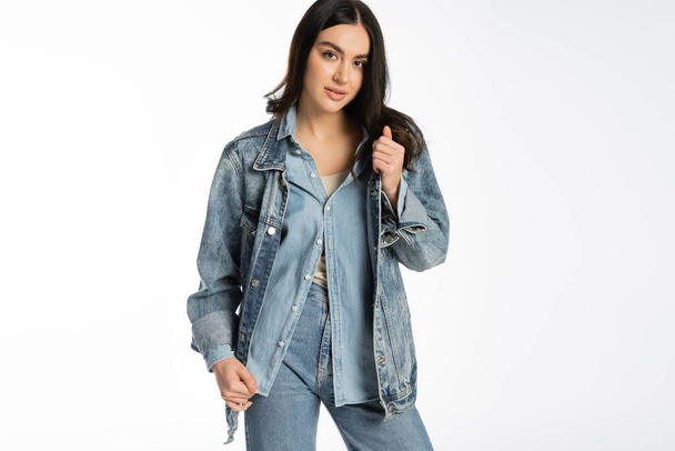 stylish young model with brunette hair and flawless makeup posing in trendy denim jacket and blue jeans while standing and looking at camera on white background  - Fotó, kép