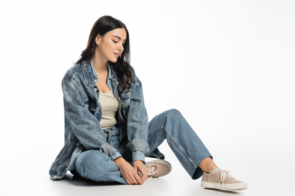 full length of alluring young woman with natural makeup and brunette hair posing in trendy denim outfit while sitting and looking away on white background - 写真・画像