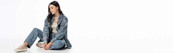 volle Länge der verführerischen Frau mit natürlichem Make-up und brünetten Haaren posiert im modischen Jeans-Outfit, während sie auf weißem Hintergrund sitzt und wegschaut, Banner  - Foto, Bild