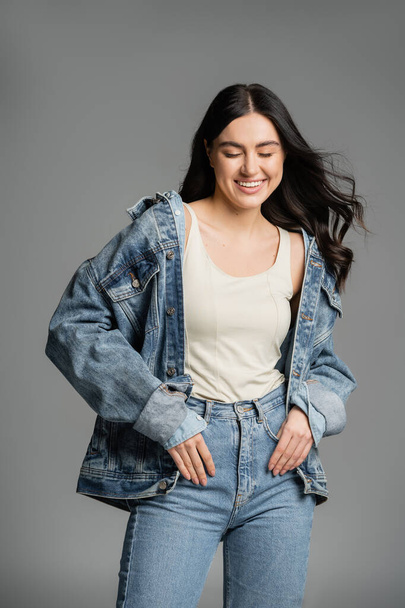 happy young woman with gorgeous brunette hair posing in stylish blue jeans and denim jacket while smiling with closed eyes on grey background  - Foto, imagen